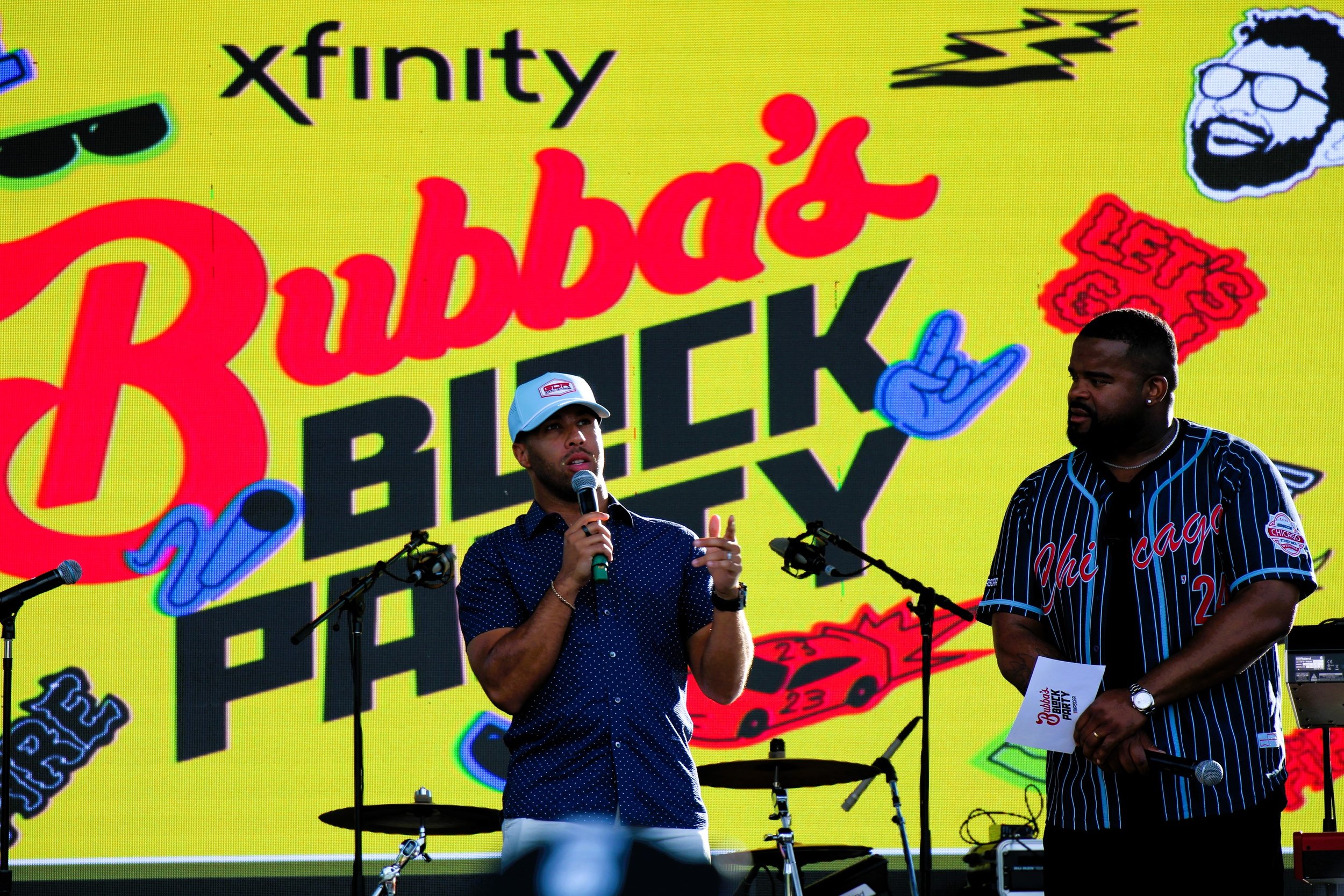 Two men on stage speaking at an event with a large colorful background featuring the logos and branding for Ruby's BBQ Party. One man holds a microphone and wears a baseball cap, while the other man holds notes and stands beside him. The backdrop displays images of cars, a person wearing sunglasses, and various decorative graphics.