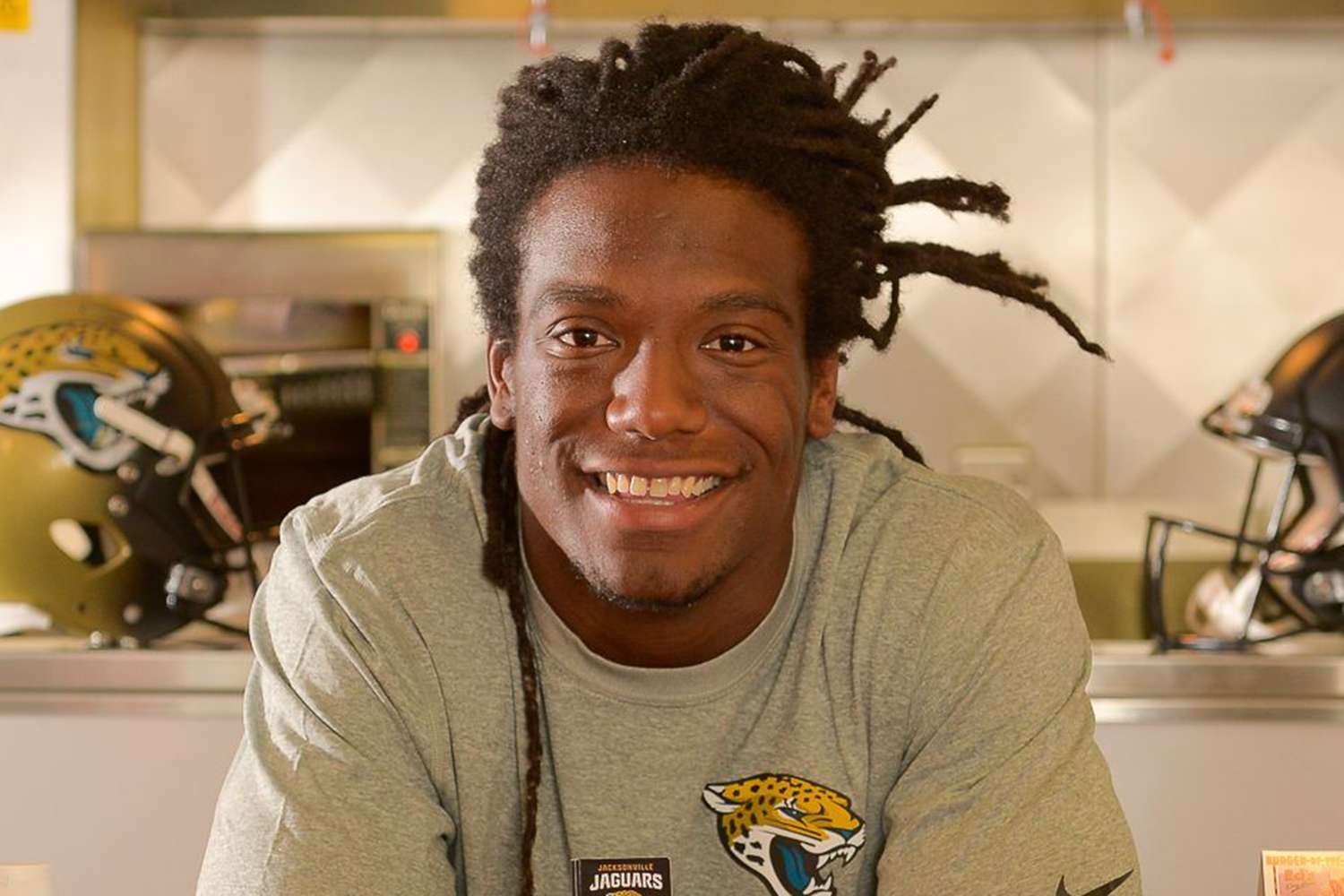 A young man with dreadlocks smiling at the camera, wearing a gray shirt with a Jacksonville Jaguars logo, with football helmets in the background.