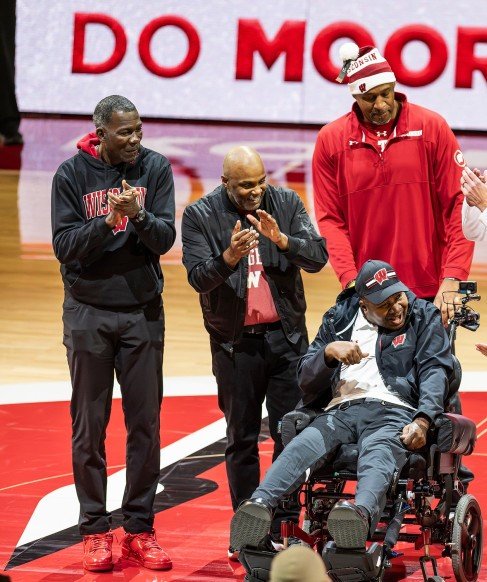 Four men on a basketball court celebrating, with one man in a wheelchair. They are smiling and clapping, wearing sports apparel, and appear to be honoring someone, with a large banner in the background.