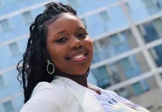 A woman with long dreadlocks, wearing a white blazer and silver hoop earrings, smiling outdoors with a modern glass building in the background.