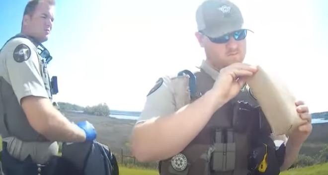 Two police officers in uniform at an outdoor location, one reading a paper bag and the other looking to the side.