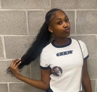 Young woman with long black hair standing against a gray brick wall, wearing a white T-shirt with navy trim and a small photo on the chest, holding her hair with one hand.