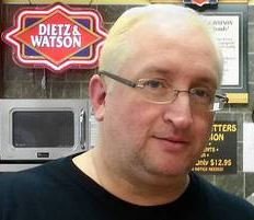 A man with glasses and light-colored hair standing in front of a Dietz Watson sign at a store.