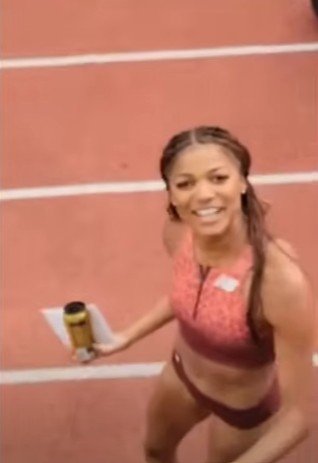 Female athlete smiling on a running track holding a water bottle and a notebook.